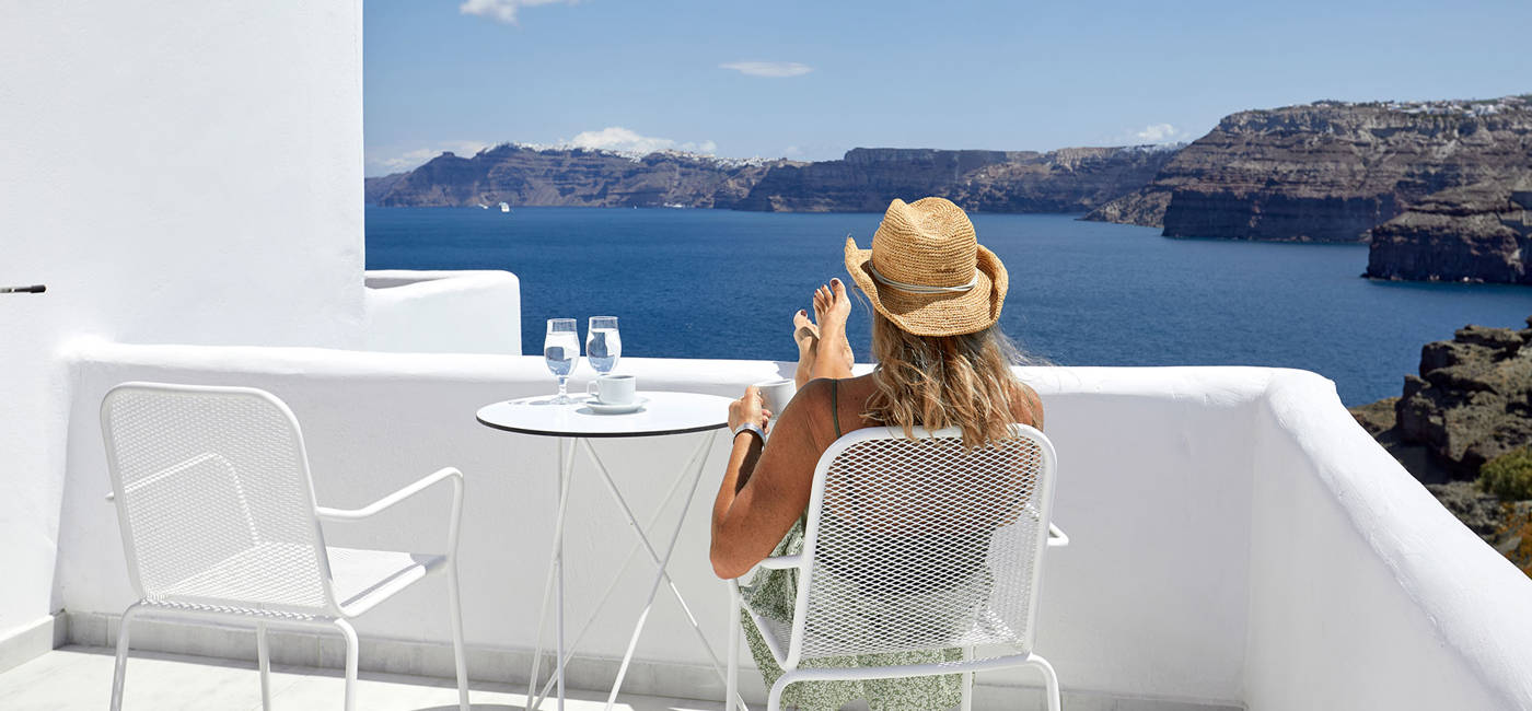 
Santorini View Hotel γυναίκα με καπέλο που κάθεται σε μπαλκόνι με θέα στη θάλασσα απολαμβάνοντας τη θέα και πίνοντας καφέ