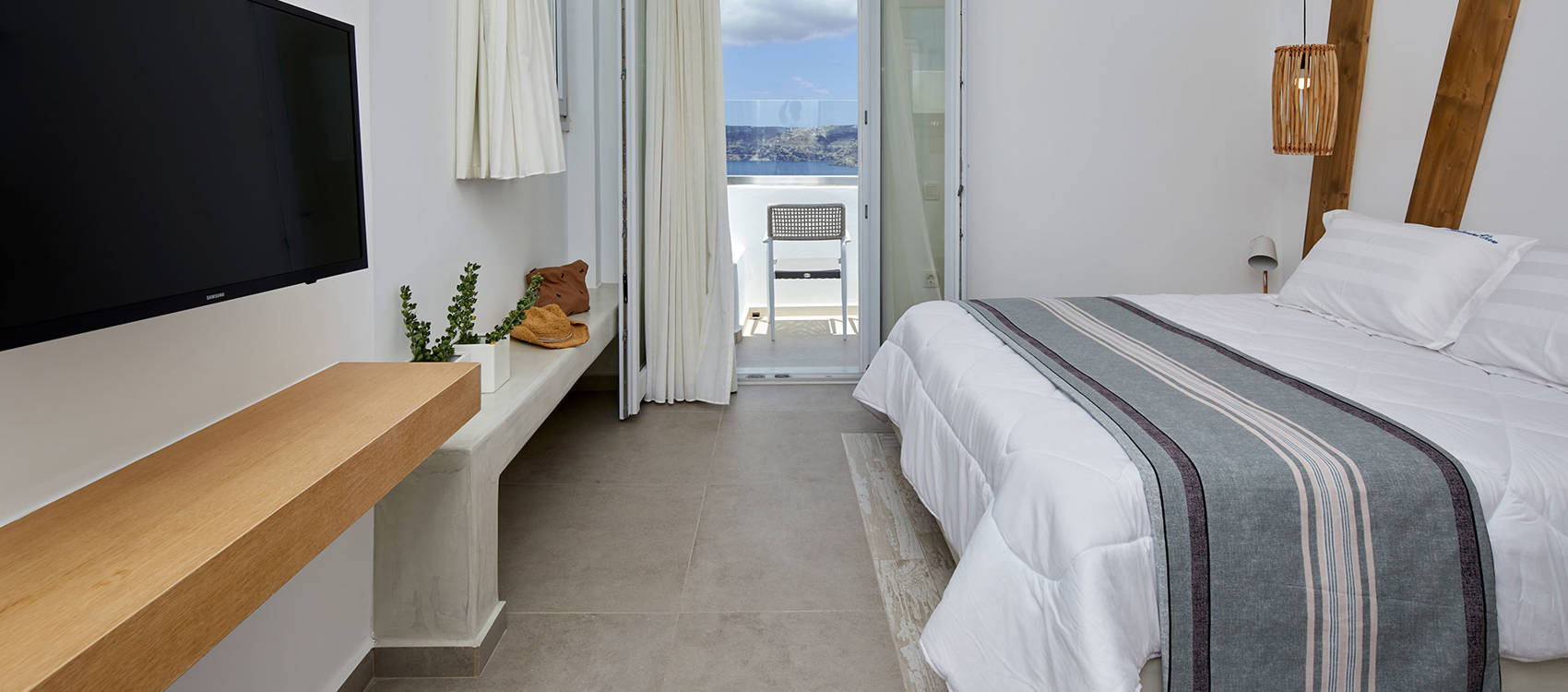  Santorini View Hotel double bed with white and grey linen, wooden decoration, white curtains and balcony with sea view