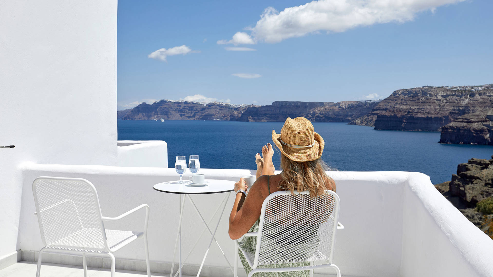 
Santorini View Hotel blonde woman with a hat seating at a sea view balcony and drinking her coffee