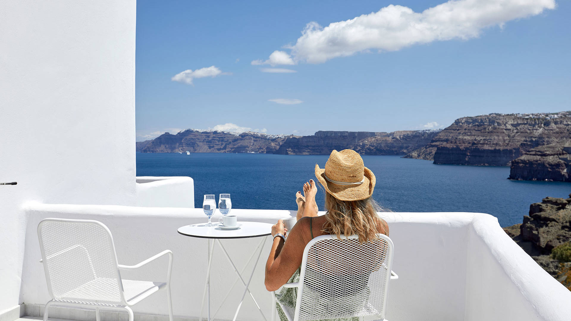
Santorini View Hotel blonde woman with a hat seating at a sea view balcony and drinking her coffee