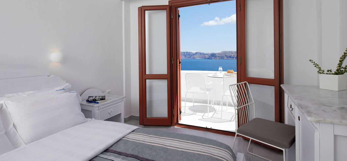 
Santorini View Hotel κρεβάτι king size και μπαλκόνι με θέα στη θάλασσα και λευκά τραπεζοκαθίσματα
